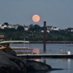 A full moon rising over the water and some boats.