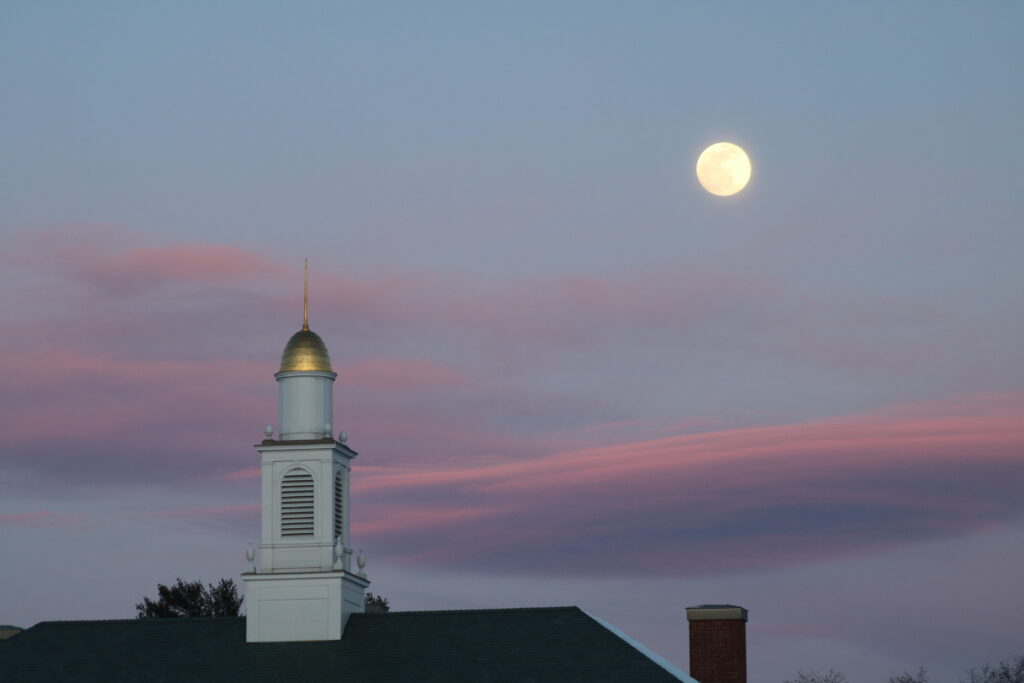 A full moon setting over the sky with a church steeple.